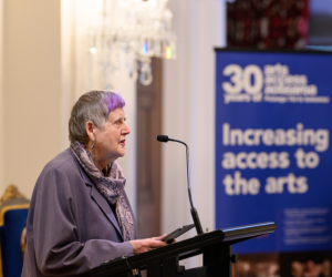 Robyn speaks into a microphone while standing at a podium, presenting a speech from her Ipad. 